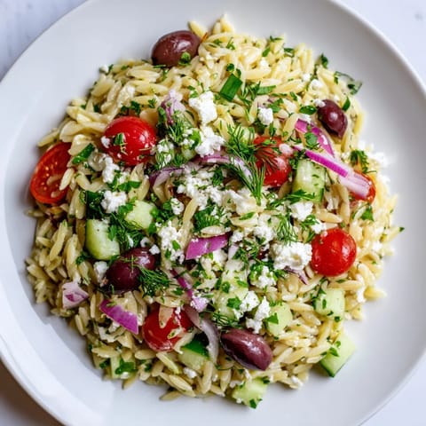 Greek Orzo Salad in a clear glass bowl showcases Kalamata olives and red onion tossed with a bright lemon-oregano dressing.