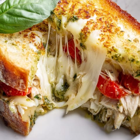 Sizzling Mozzarella Pesto Chicken Grilled Cheese with golden sourdough, melted cheese, and fresh basil on a plate.  